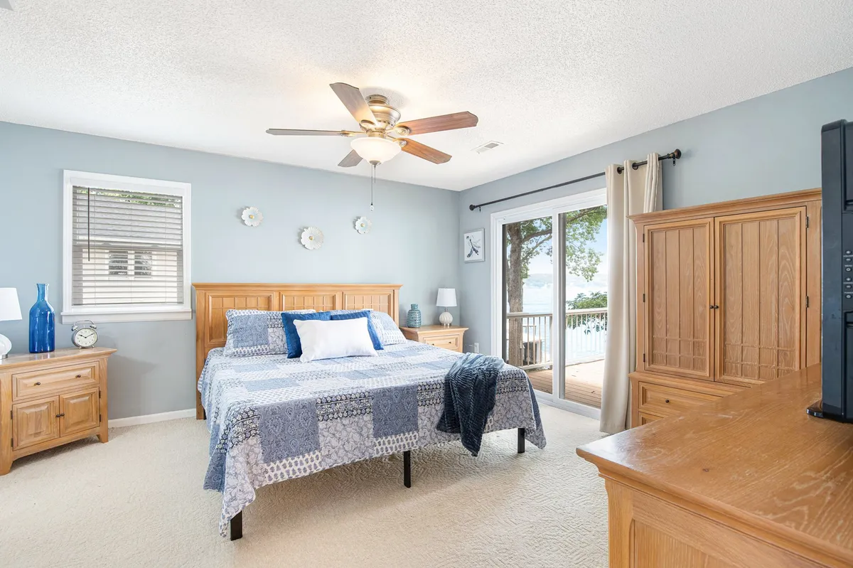 A tranquil bedroom featuring a neatly made bed with a colorful quilt, large windows letting in natural light, a functional dresser, and a bedside lamp. The view through the windows hints at a peaceful outdoor setting, creating a serene atmosphere perfect for relaxation.