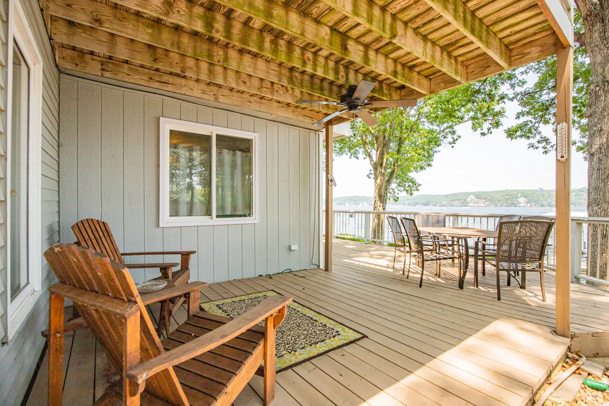 An outdoor deck setup with wooden chairs and a dining table facing out towards the lake, with trees in the background. The scene is tranquil and designed for open-air meals, relaxation, and group gatherings, especially during sunset.