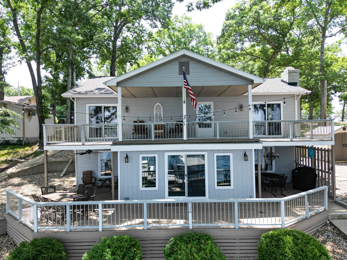 A spacious wooden deck bordered by lush greenery, featuring multiple seating areas under an open sky. The American flag flutters while the deck invites gatherings with tables and chairs arrayed for outdoor dining and relaxation. The layout hints at ample space for games, meals, or sunset viewing.