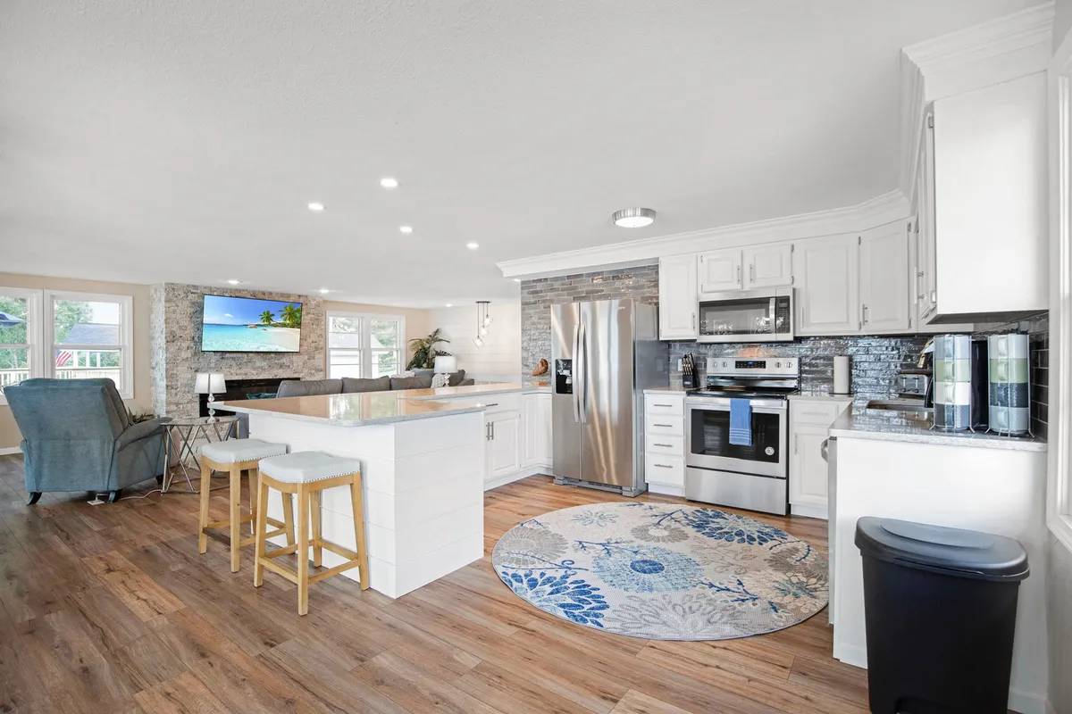A bright, spacious kitchen with a central island and bar stools, stainless steel appliances, a decorative rug, and large windows that flood the space with natural light. A television is mounted on the wall, making the space feel modern and multi-functional.