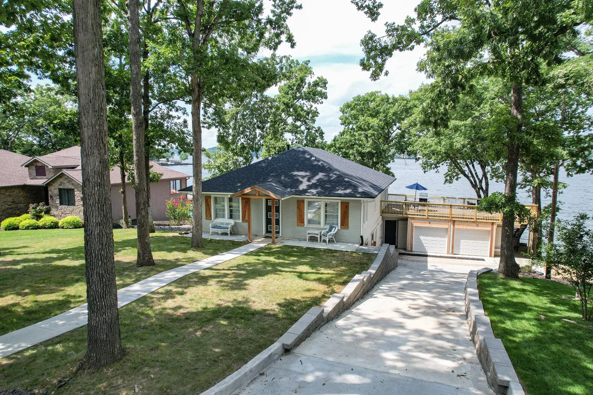 A peaceful exterior view of one of the homes, showcasing the house, driveway, deck by the water, and surrounding trees. The ample driveway space illustrates the convenience of parking for larger groups, while the deck and yard highlight prime spots for shared celebrations and lakeside relaxation.