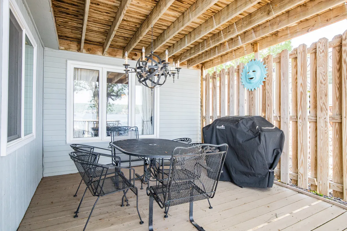 A cozy outdoor dining area featuring a table with chairs, warm lighting from a chandelier, a barbecue grill, and a decorative sun motif on the wall. The setup signals a space designed for communal meals, barbecues, and socializing into the evening.