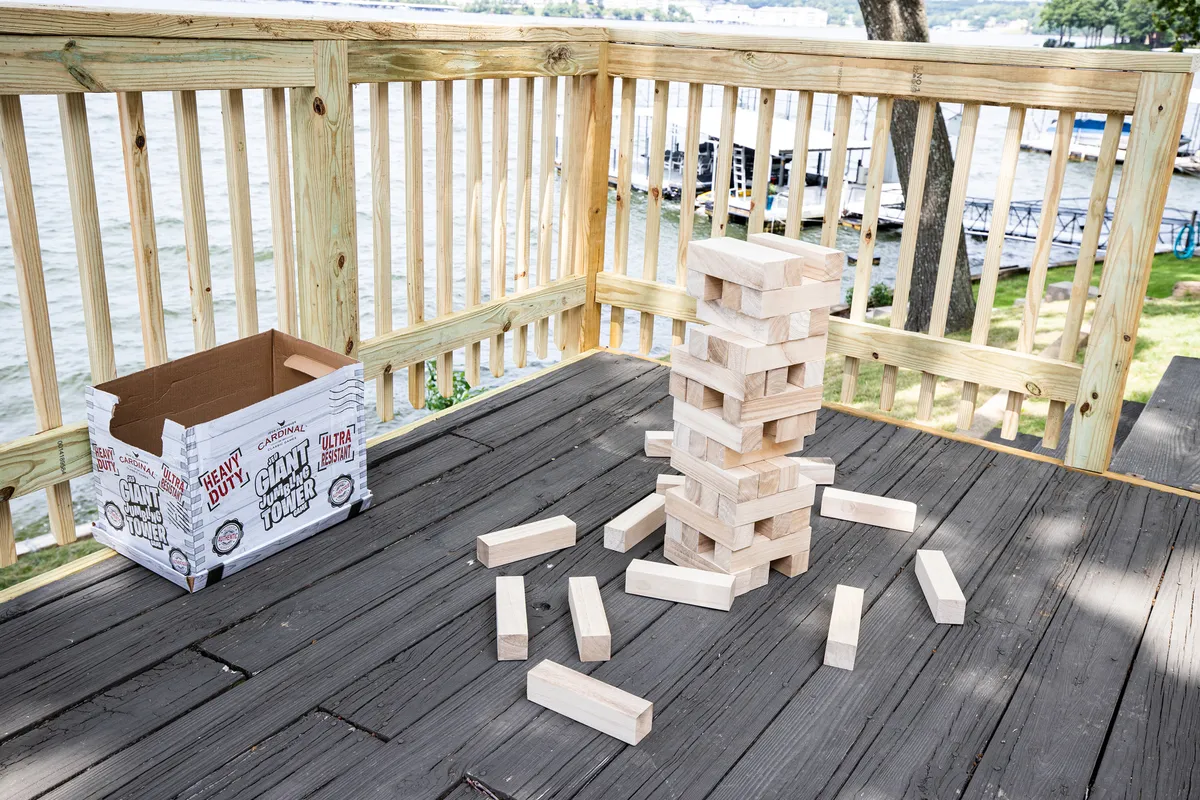 A lively outdoor scene focused on a giant Jenga game set atop a deck, with scenic views in the background. The image captures the energy and fun of group games, emphasizing interactive entertainment and shared laughter during gatherings.