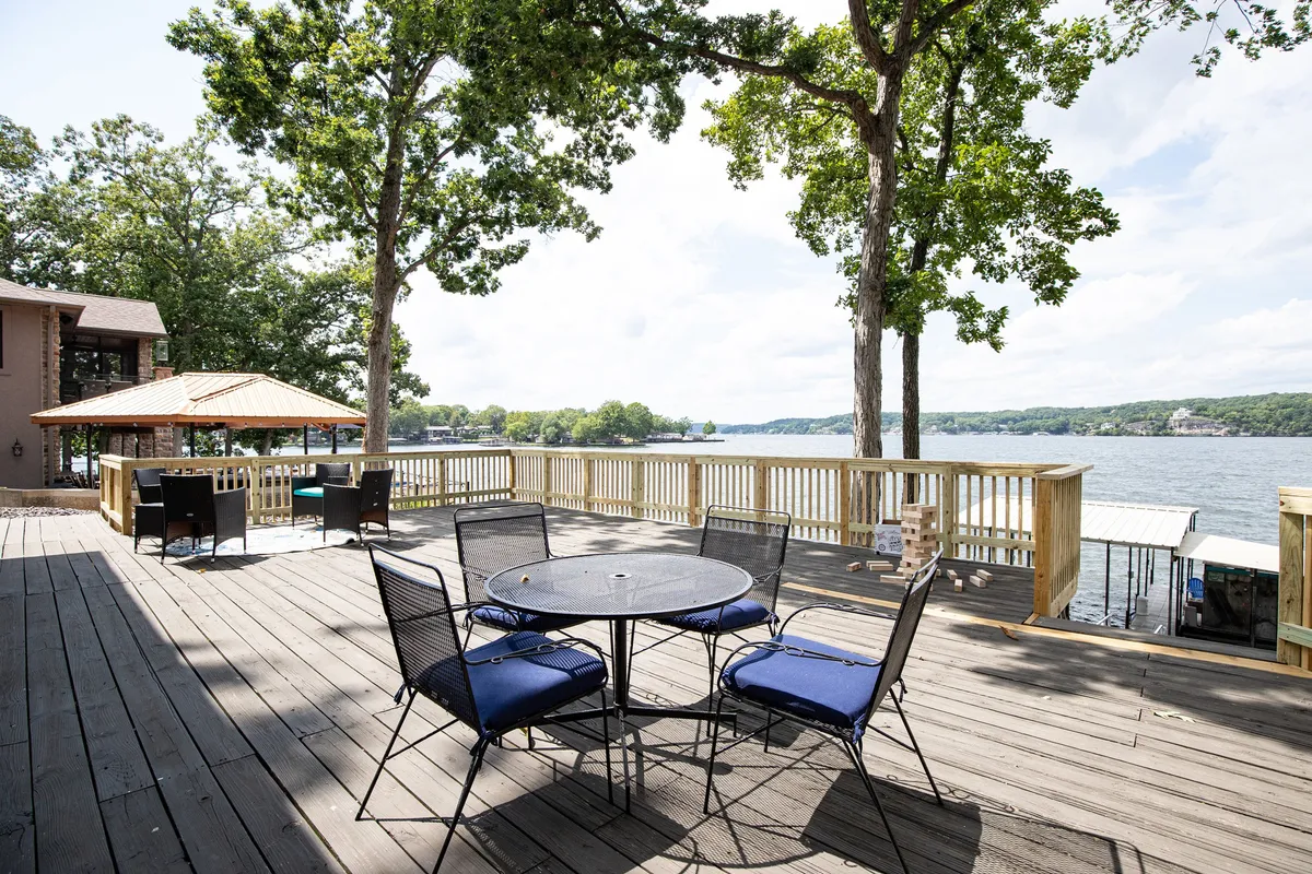 A spacious deck with a round table and blue chairs overlooks the lake and dock, framed by trees. The area is set for outdoor dining, relaxing, or enjoying games while taking in the water views.