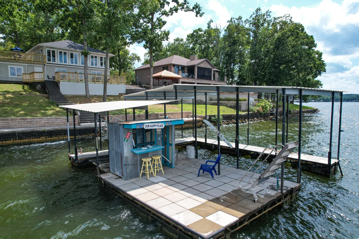 A cheerful dock scene featuring a blue bar with the sign 'OUR HAPPY PLACE,' stools, a lounge chair, and direct lake access. The dock is equipped for various water activities and is perfect for relaxing by the water or launching boats and jet skis.