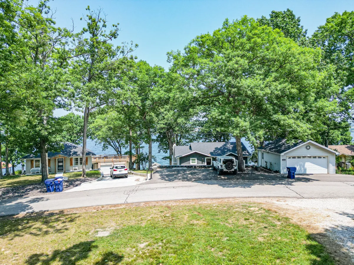 A scenic view of large green trees surrounding a peaceful lake, with nearby houses and a driveway with parked cars visible in the foreground. The lake in the background suggests easy access to water activities and the outdoors.